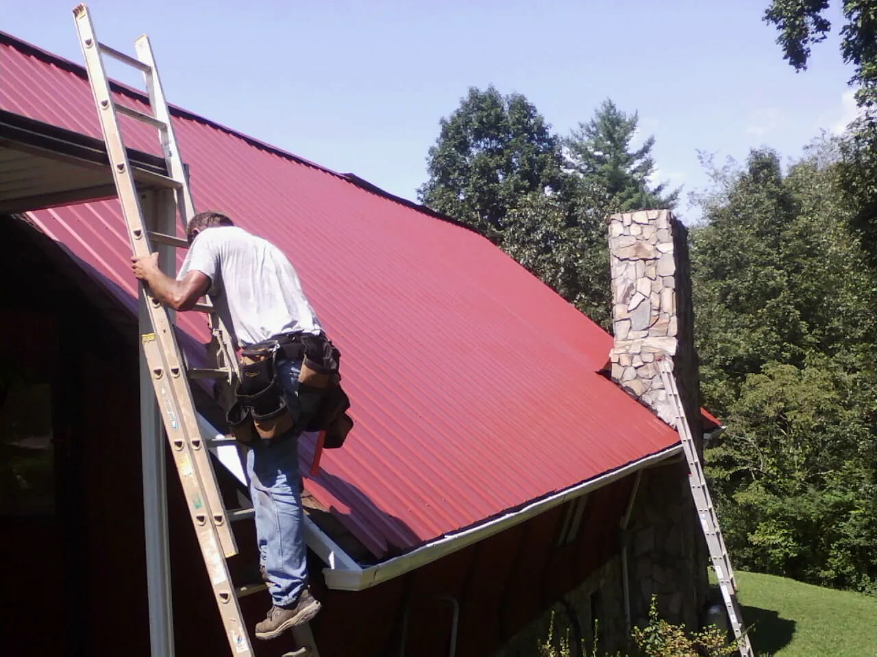 Roofer on a ladder beside a red metal roof and stone chimney — Soffit & Fascia Repair by Roofing Brothers in Jacobsburg